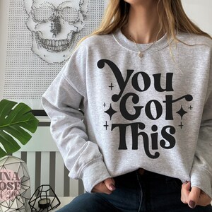 Puede incluir: Una sudadera gris con el texto "You Got This" en negro, en letras estilizadas. El texto est&aacute; rodeado de peque&ntilde;os signos m&aacute;s y diamantes.