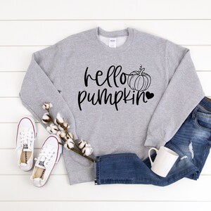 May include: A gray sweatshirt with the text "hello pumpkin" in black lettering and a black outline of a pumpkin with a heart. The sweatshirt is laying on a white wooden surface with a pair of white sneakers and a pair of blue jeans.