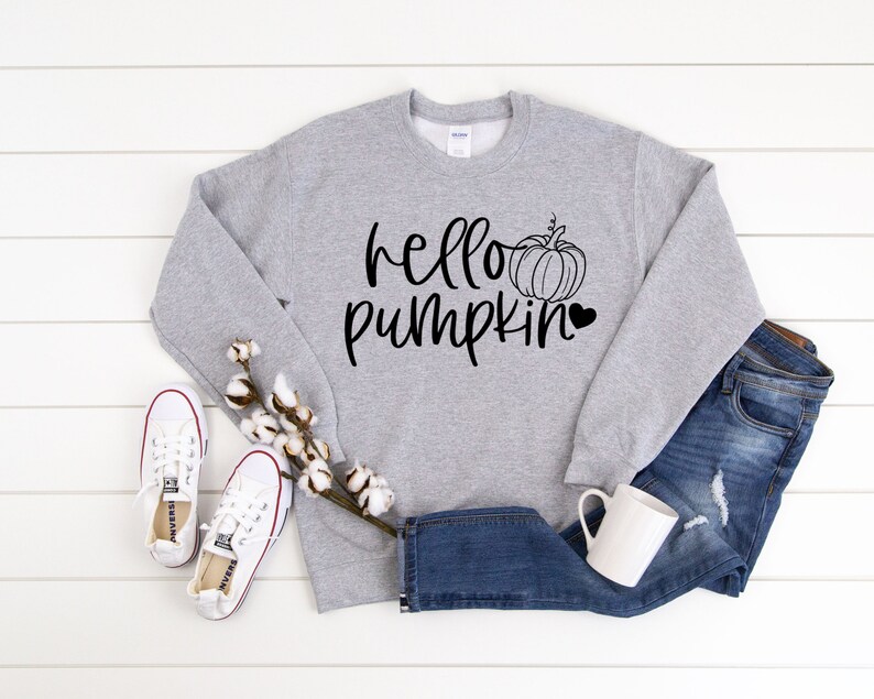 May include: A gray sweatshirt with the text "hello pumpkin" in black lettering and a black outline of a pumpkin with a heart. The sweatshirt is laying on a white wooden surface with a pair of white sneakers and a pair of blue jeans.