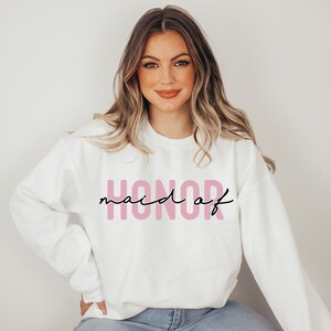 May include: White sweatshirt with the text "maid of honor" in pink letters and black script.