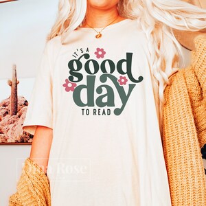 Op de afbeelding: Een wit T-shirt met de tekst "It's a good day to read" in groen en roze. De tekst is omringd door roze bloemen.