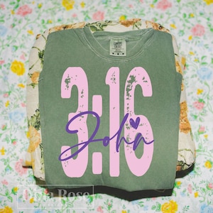 May include: An olive green Comfort Colors sweatshirt with the text "3:16 John" in pink, with a small purple heart. The sweatshirt is folded and resting on a floral patterned fabric.