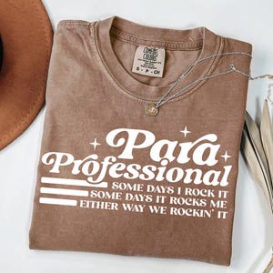 May include: A brown t-shirt with the words "Para Professional" in white script. The shirt also has the text "SOME DAYS I ROCK IT, SOME DAYS IT ROCKS ME, EITHER WAY WE ROCKIN' IT". A silver necklace with a pendant is on the shirt.