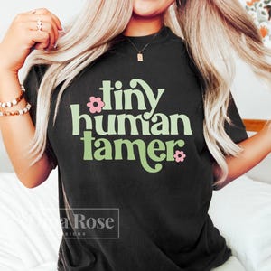 May include: A black t-shirt with a green and pink graphic that says "tiny human tamer".