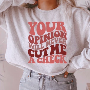 May include: A gray sweatshirt with the text "YOUR OPINION WILL NEVER CUT ME A CHECK" in a retro, colorful font.