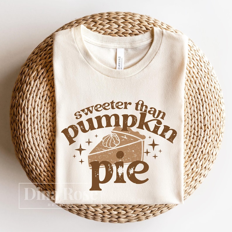 Pode incluir: Uma camiseta bege com o texto "sweeter than pumpkin pie" em letras marrons. O texto est&aacute; em uma fonte de estilo vintage e inclui um gr&aacute;fico de uma fatia de torta de ab&oacute;bora com chantilly.