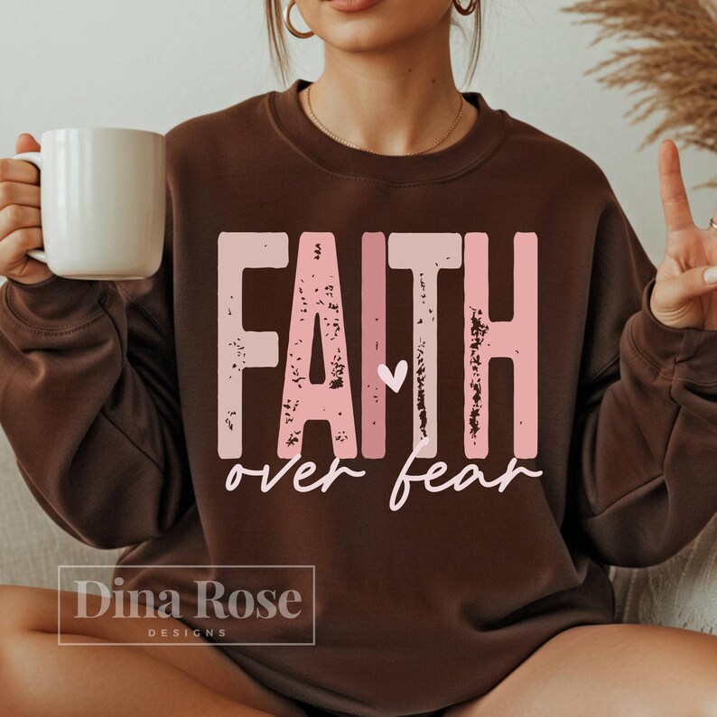 May include: A brown sweatshirt with the words "FAITH over fear" in a distressed pink font. A small white heart is between the words. The brand name "Dina Rose Designs" is at the bottom.