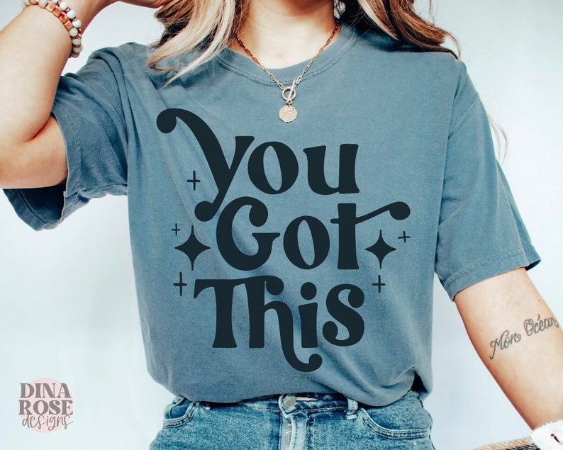 Puede incluir: Una camiseta azul con el texto "You Got This" en una fuente retro. El texto est&aacute; en negro y est&aacute; rodeado de peque&ntilde;as estrellas.