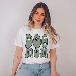 May include: White t-shirt with a green "Dog Mom" graphic featuring white flowers. The graphic is in a retro style.