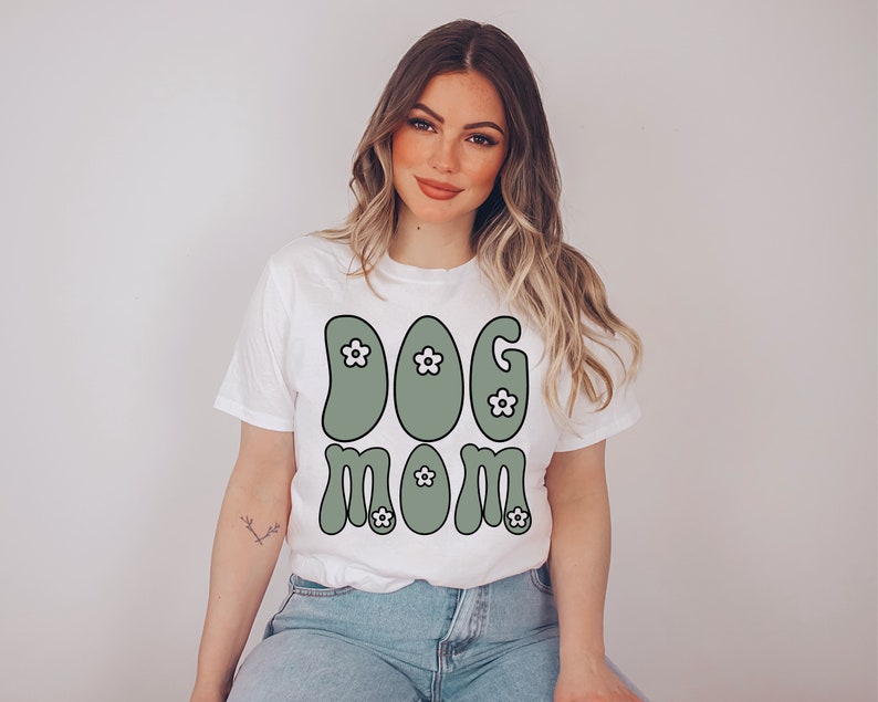 May include: White t-shirt with a green "Dog Mom" graphic featuring white flowers. The graphic is in a retro style.