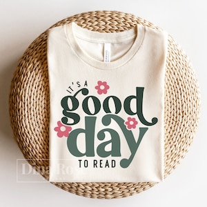 Op de afbeelding: Een beige T-shirt met de tekst "It's a good day to read" in groen en roze. De tekst is gestyled met een bloemenontwerp.