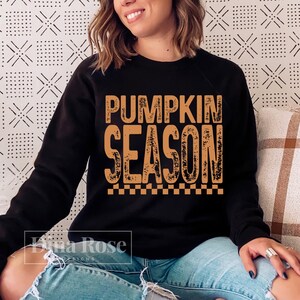 May include: Black sweatshirt with the text "PUMPKIN SEASON" in orange, with a checkered pattern at the bottom. The sweatshirt is worn by a person. The background is a patterned white and black design.