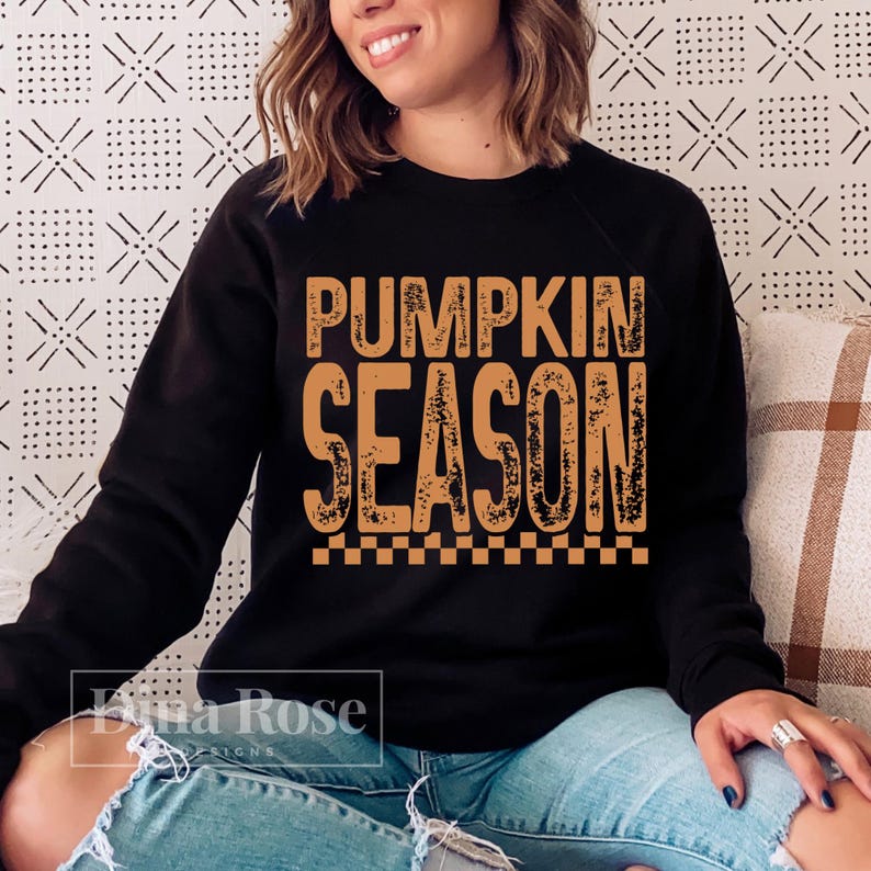 May include: Black sweatshirt with the text "PUMPKIN SEASON" in orange, with a checkered pattern at the bottom. The sweatshirt is worn by a person. The background is a patterned white and black design.