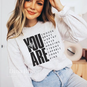 May include: A white sweatshirt with the words "YOU ARE" in large black letters. The letters are surrounded by smaller black words that spell out "enough, brave, worthy, loved, amazing, strong, capable, smart, kind".