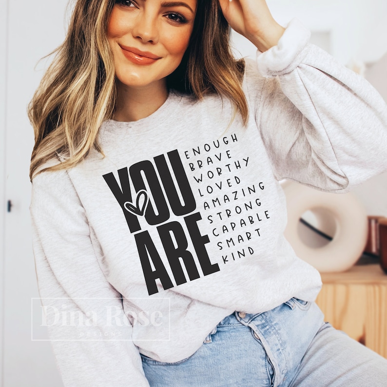 May include: A white sweatshirt with the words "YOU ARE" in large black letters. The letters are surrounded by smaller black words that spell out "enough, brave, worthy, loved, amazing, strong, capable, smart, kind".