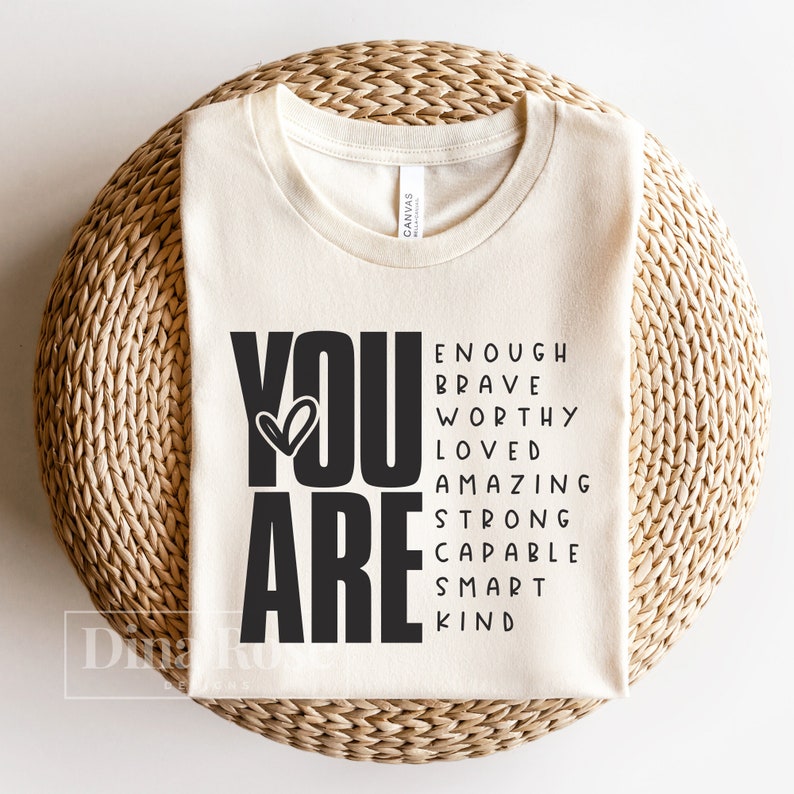May include: A beige t-shirt with the words "YOU ARE" in black, with a heart symbol in the "O". The words "ENOUGH, BRAVE, WORTHY, LOVED, AMAZING, STRONG, CAPABLE, SMART, KIND" are arranged around the words "YOU ARE".