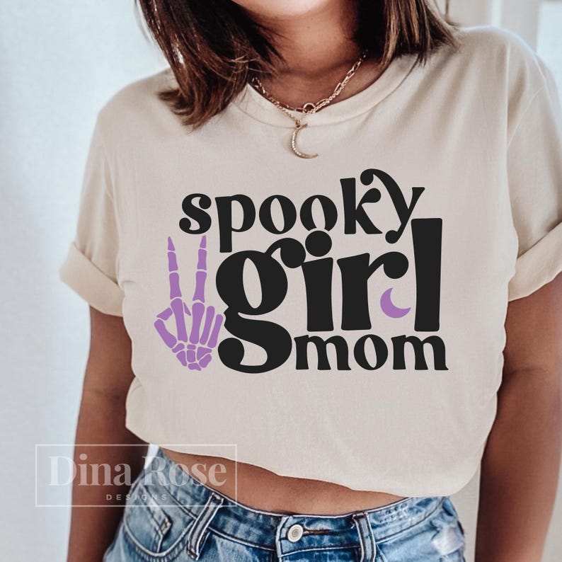 May include: A white t-shirt with the text "spooky girl mom" in black lettering. A purple skeleton hand with a peace sign is to the left of the text.