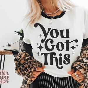 Puede incluir: Camiseta blanca con texto negro que dice "You Got This" en una fuente retro. El texto est&aacute; rodeado de peque&ntilde;as estrellas negras.