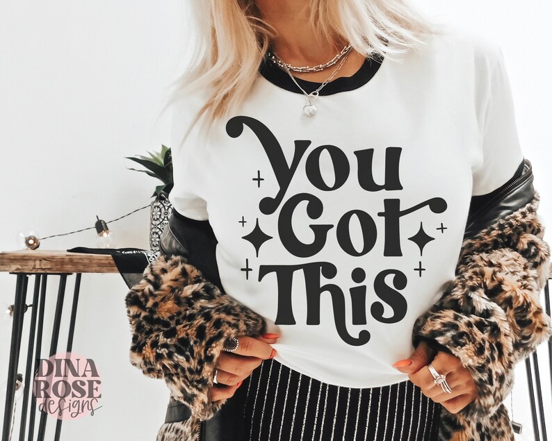 Puede incluir: Camiseta blanca con texto negro que dice "You Got This" en una fuente retro. El texto est&aacute; rodeado de peque&ntilde;as estrellas negras.