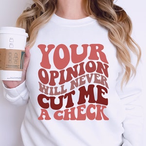 May include: White sweatshirt with the text "YOUR OPINION WILL NEVER CUT ME A CHECK" in a retro font with pink, red, and brown colors.