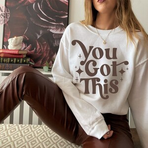 Puede incluir: Sudadera blanca con texto marr&oacute;n que dice "You Got This" en un estilo de fuente retro.