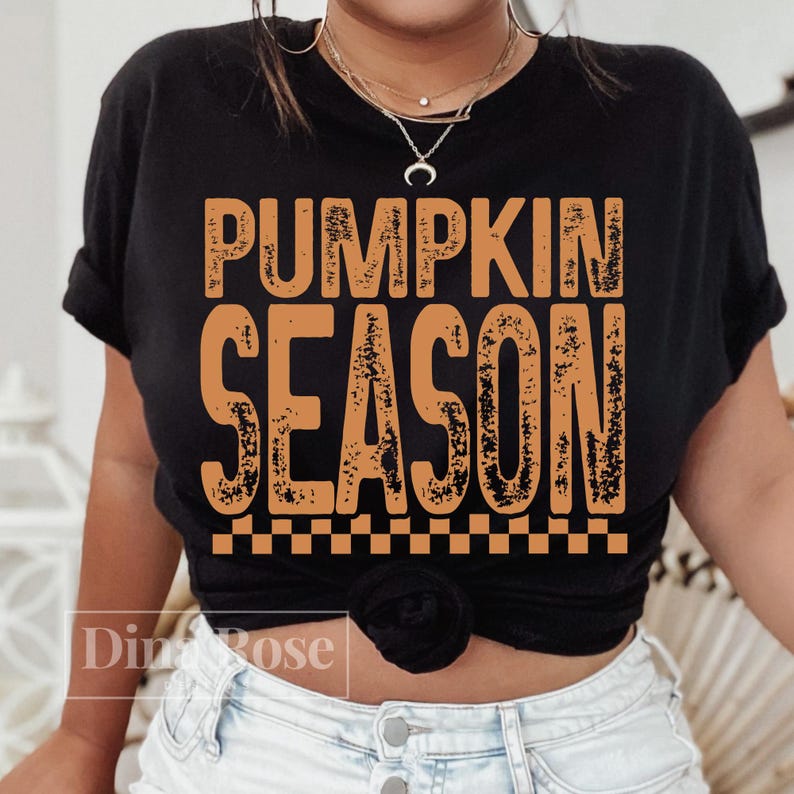 May include: Black t-shirt with the words "PUMPKIN SEASON" in orange, distressed lettering. The shirt has a checkered pattern at the bottom of the text. The shirt is worn by a person.