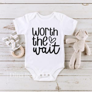 May include: White baby onesie with black text that reads "Worth the wait" with a small heart symbol.