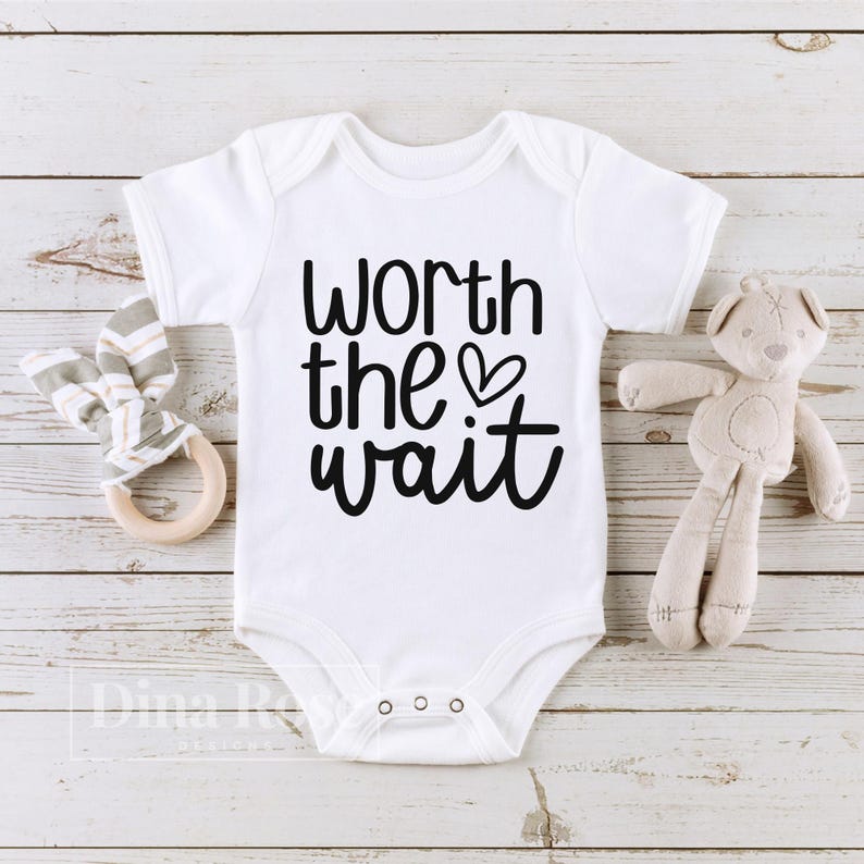 May include: White baby onesie with black text that reads "Worth the wait" with a small heart symbol.
