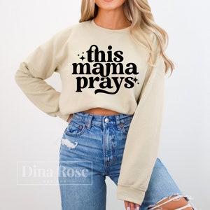 May include: A beige cropped sweatshirt with the text "this mama prays" in black retro-style lettering.
