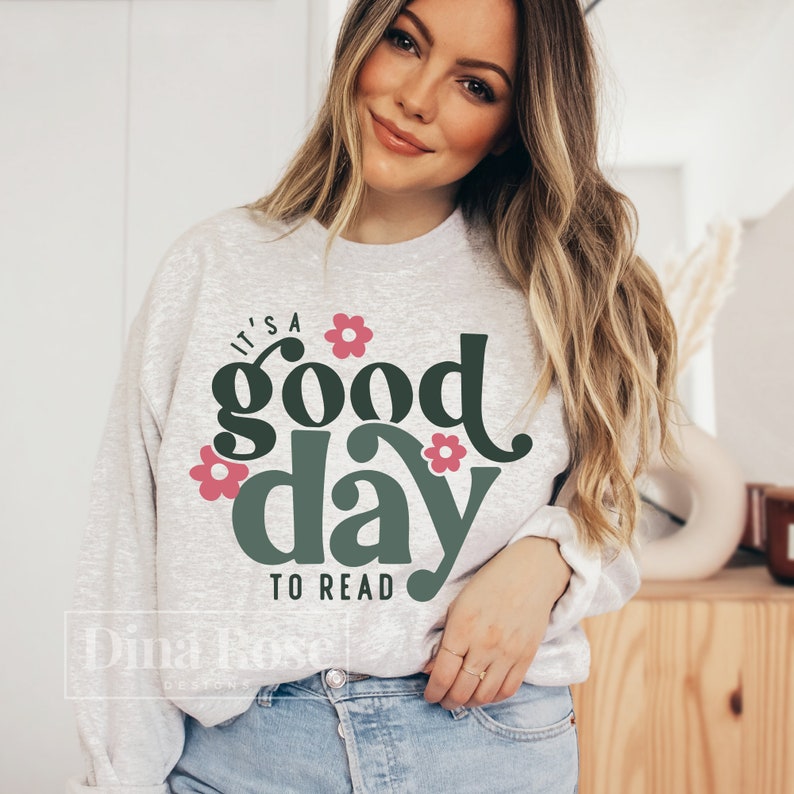 Op de afbeelding: Een grijs sweatshirt met de tekst "It's a good day to read" in groen en roze. De tekst is omringd door roze bloemen.