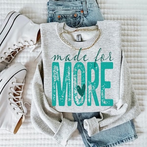 K&ouml;nnte beinhalten: Ein hellgraues Sweatshirt mit den Worten "made for MORE" in t&uuml;rkisfarbener Schrift und Blockbuchstaben, mit einem t&uuml;rkisfarbenen Herz. Das Sweatshirt wird mit einer blauen Jeans, einer goldenen Halskette und wei&szlig;en Turnschuhen kombiniert.