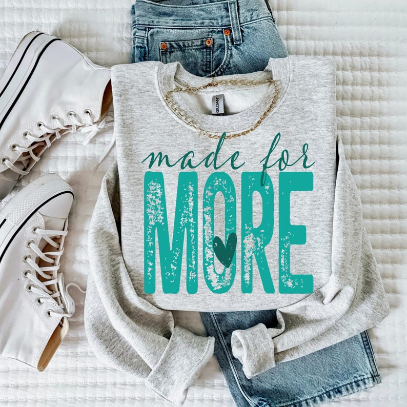 K&ouml;nnte beinhalten: Ein hellgraues Sweatshirt mit den Worten "made for MORE" in t&uuml;rkisfarbener Schrift und Blockbuchstaben, mit einem t&uuml;rkisfarbenen Herz. Das Sweatshirt wird mit einer blauen Jeans, einer goldenen Halskette und wei&szlig;en Turnschuhen kombiniert.