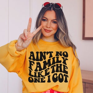 Pu&ograve; includere: Una felpa gialla con testo nero che recita "Ain't no family like the one I got" in un font retr&ograve;.
