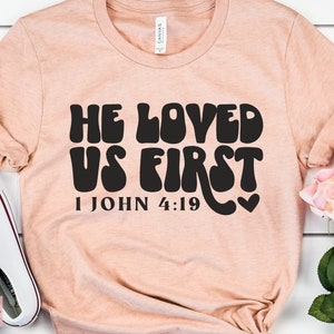 May include: A peach-colored t-shirt with the text "He Loved Us First" in black, stylized lettering. The text "1 John 4:19" is also printed in black below the main text.