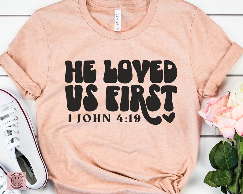 May include: A peach-colored t-shirt with the text "He Loved Us First" in black, stylized lettering. The text "1 John 4:19" is also printed in black below the main text.