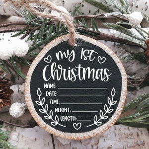 Pu&ograve; includere: Un ornamento rotondo in legno con una superficie in lavagna nera. Dice "my 1st Christmas" con cuori e uno spazio per scrivere il nome, la data, l'ora, il peso e la lunghezza del bambino.