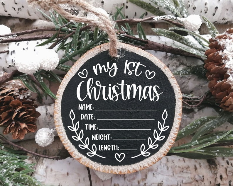 Pu&ograve; includere: Un ornamento rotondo in legno con una superficie in lavagna nera. Dice "my 1st Christmas" con cuori e uno spazio per scrivere il nome, la data, l'ora, il peso e la lunghezza del bambino.