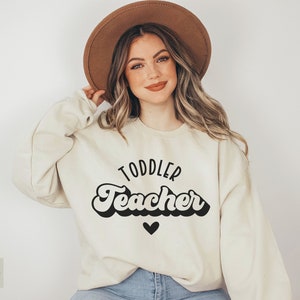 Pu&ograve; includere: Una donna indossa una felpa color crema con la scritta "Toddler Teacher" in un font retr&ograve;. Il testo &egrave; nero e sotto c'&egrave; un piccolo cuore nero.