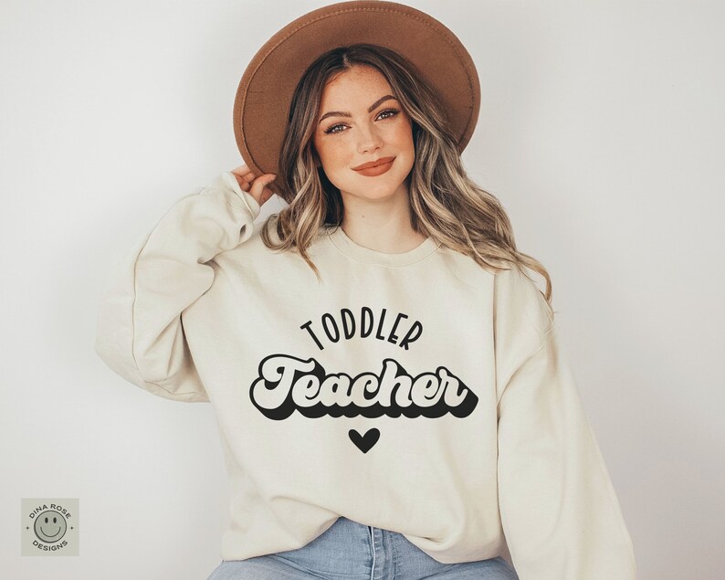 Pu&ograve; includere: Una donna indossa una felpa color crema con la scritta "Toddler Teacher" in un font retr&ograve;. Il testo &egrave; nero e sotto c'&egrave; un piccolo cuore nero.