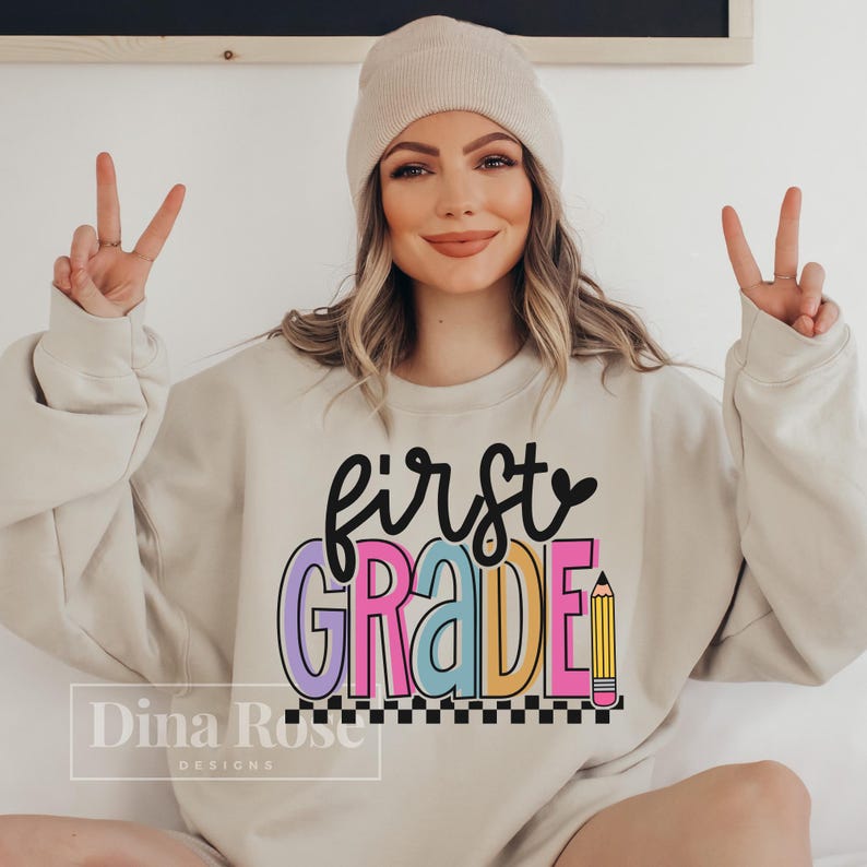 K&ouml;nnte beinhalten: Beiges Sweatshirt mit den Worten "first grade" in schwarzer Schrift und bunten Blockbuchstaben. Eine gelbe Bleistiftgrafik befindet sich neben dem Wort "grade". Das Sweatshirt hat einen schwarz-wei&szlig;en karierten Rand am unteren Rand.