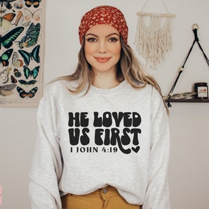 May include: A gray sweatshirt with the text "He Loved Us First" in black, with a retro font style. The text is accompanied by the scripture reference "1 John 4:19" and a small heart symbol.