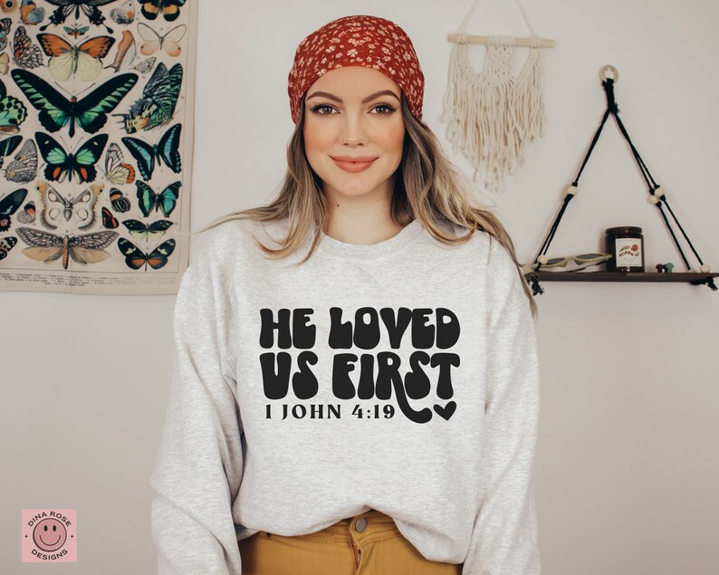 May include: A gray sweatshirt with the text "He Loved Us First" in black, with a retro font style. The text is accompanied by the scripture reference "1 John 4:19" and a small heart symbol.