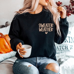 May include: Black sweatshirt with the text "SWEATER WEATHER" in a light brown font, with a small heart below the text. The sweatshirt is being worn by a person holding a white mug. The person is wearing blue jeans.