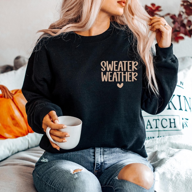May include: Black sweatshirt with the text "SWEATER WEATHER" in a light brown font, with a small heart below the text. The sweatshirt is being worn by a person holding a white mug. The person is wearing blue jeans.