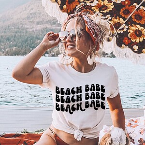 May include: A white t-shirt with the text "Beach Babe" repeated three times in a retro, wavy font. The shirt is worn by a person with a red bandana in their hair and sunglasses.