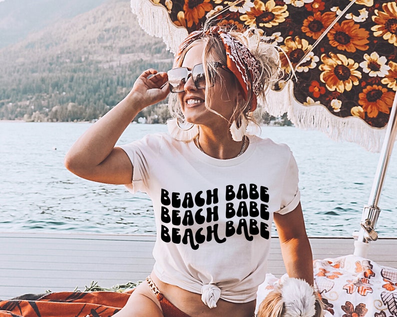 May include: A white t-shirt with the text "Beach Babe" repeated three times in a retro, wavy font. The shirt is worn by a person with a red bandana in their hair and sunglasses.