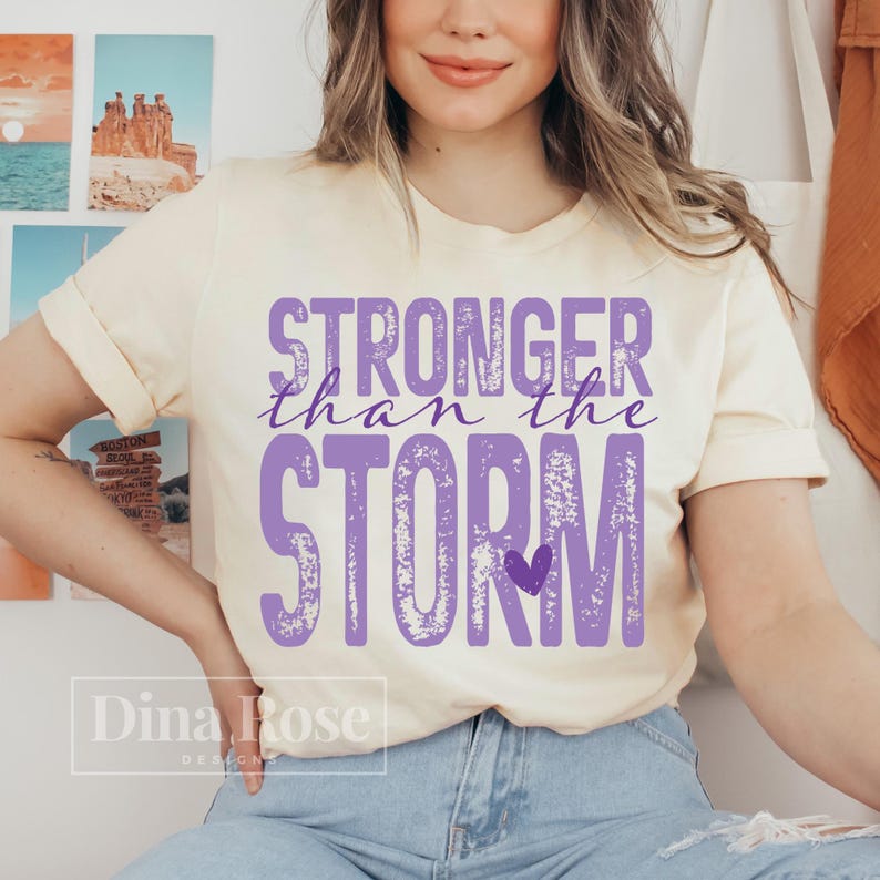 K&ouml;nnte beinhalten: Cremefarbenes T-Shirt mit dem Text "STRONGER than the STORM" in Lila. Unter dem Text befindet sich ein kleines lila Herz. Das Shirt ist ein l&auml;ssiger Stil.