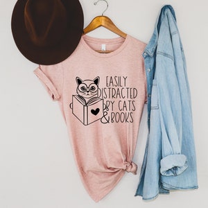 K&ouml;nnte beinhalten: Ein rosa T-Shirt mit einer schwarzen Grafik eines Katzen mit Brille, das ein Buch liest. Der Text auf dem Shirt lautet "Easily Distracted by Cats & Books".