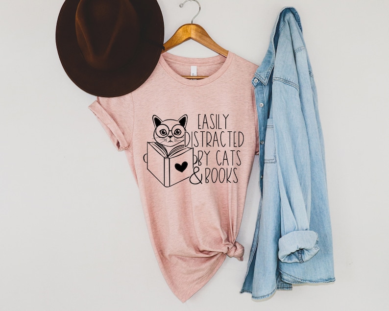 K&ouml;nnte beinhalten: Ein rosa T-Shirt mit einer schwarzen Grafik eines Katzen mit Brille, das ein Buch liest. Der Text auf dem Shirt lautet "Easily Distracted by Cats & Books".