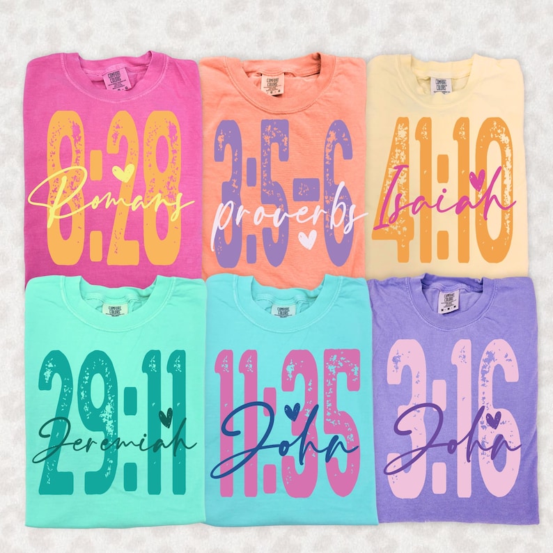 May include: Six colorful t-shirts are displayed, each with a unique design. The shirts feature large numbers and script text, including the words "Romans," "Proverbs," "Isaiah," "Jeremiah," and "John." Each shirt has a different color, including pink, orange, yellow, teal, blue, and purple.